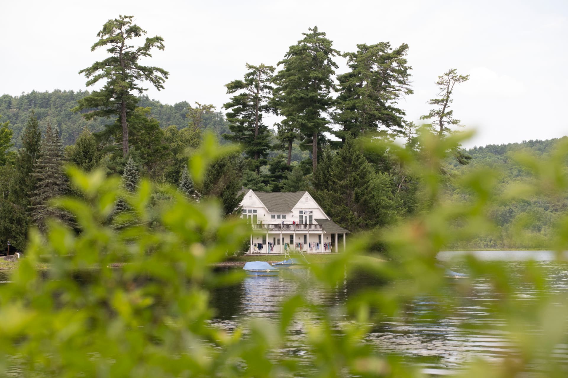 The white boathouse on Brant Lake seen through pine branches, with tall trees behind